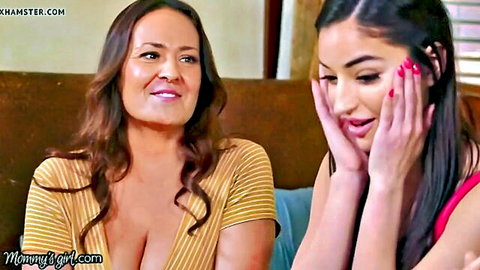 Brunette MILF with deep cleavage smiles at surprised young Latina covering mouth on couch