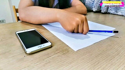 Southeast Asian teen girl in uniform writing on paper with pencil next to phone at desk