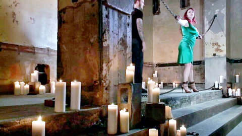 Redhead dominatrix in tight green dress holds chain on chained submissive man in candlelit BDSM dungeon