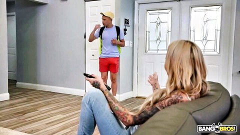 Tattooed blonde MILF with big tits on couch greets young guy in red shorts and backpack
