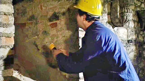 Worker in yellow hardhat and blue overalls plastering rough stone wall with trowel
