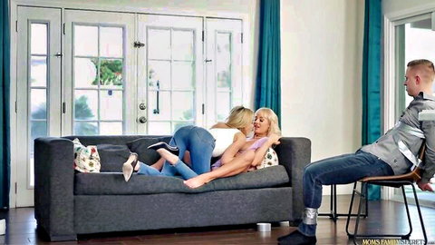Two blonde MILFs in jeans and heels lounge intimately on couch, man watches