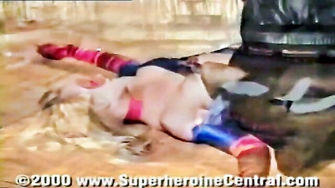 Blonde Ultrawoman in red and blue costume lying defeated on sand in peril trap scene