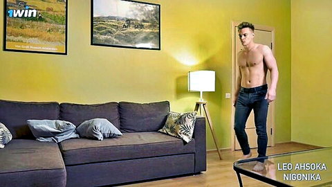 Shirtless muscular hunk in tight jeans stands in yellow-walled living room