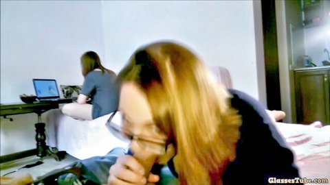 Blonde woman with glasses sucks toy on bed, legs spread, friend at laptop
