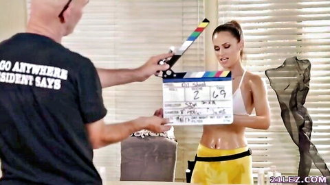 Tanned Latina in skimpy white bikini top and yellow bottoms on lesbian porn set with director clapperboard