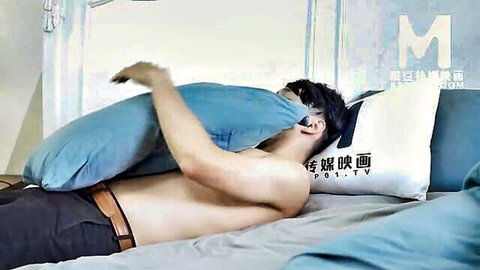 Shirtless fit Asian man lying in bed, hiding face with blue pillow, teasing pose