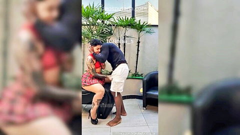 Ebony man embraces tattooed busty babe in red top and plaid skirt from behind outdoors