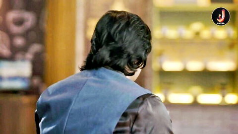 Back view of long-haired South Asian man in blue vest standing in front of golden shelves in romantic Indian episode