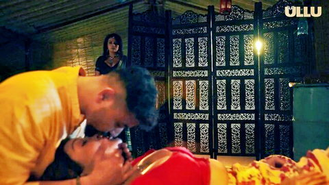 Indian man passionately kissing busty wife's neck in bed as voyeur woman peeks from screen