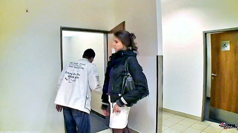 Brunette in short skirt stands as doctor in lab coat bends down between her legs in public hallway