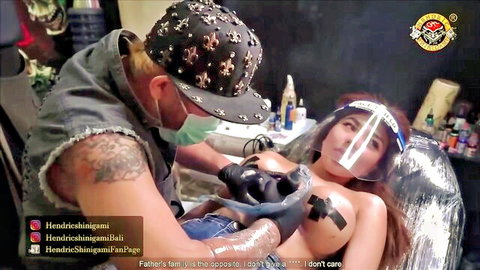 Tattoo artist gloves up to ink topless woman's bare tits in Bali parlor