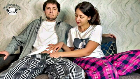Smiling brunette teen in pink plaid pajamas holds guy's hand on her thigh during playful couch cuddle