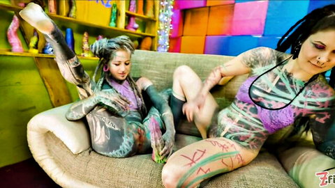 Tattooed dreadlock lesbians spreading legs fingering pussies on couch amid colorful dildos