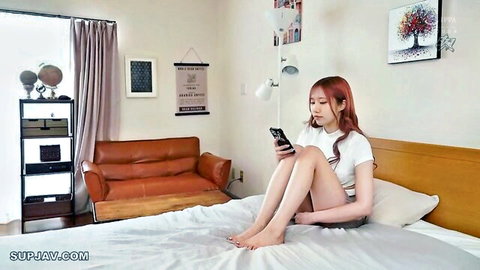 Redhead East Asian babe in crop top and tiny shorts sits legs spread on bed holding phone
