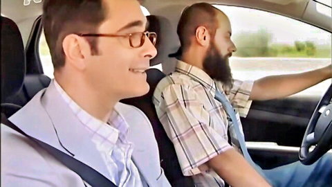 Two Greek men in car, bearded driver in suspenders, suited passenger smiling