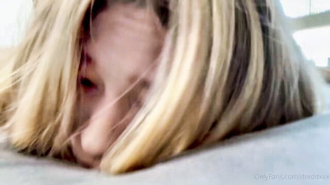 POV closeup of blonde babe's face with long hair spread on bed, mouth open ready to suck cock