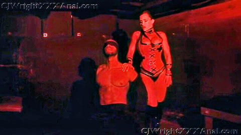 Tanned curvy dominatrix in red latex corset leashes blindfolded muscular man in BDSM dungeon