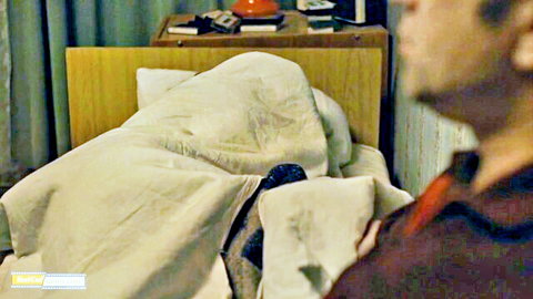 Latina in dorm bed hiding under white sheets, man watching from side