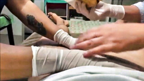 Gloved Asian masseuse wrapping white bandages on young woman's bare legs during erotic massage