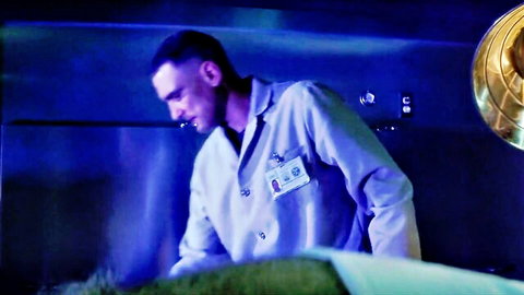 Mysterious man in white lab coat standing in blue-lit room over exam table
