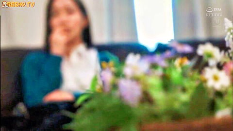 Blurry Asian woman with long hair in teal top posing thoughtfully behind colorful flowers