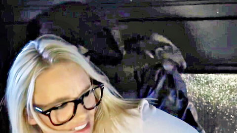Blonde babe in glasses smiles seductively in dark car with shadowy figure behind her