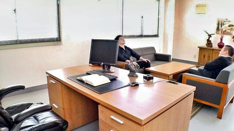 Asian office woman in black suit reclining relaxed on couch watched by suited man