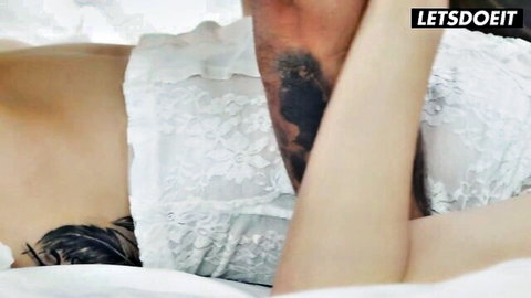 Tattooed stud holds up big ass brunette babe's leg exposing shaved pussy on lace sheets