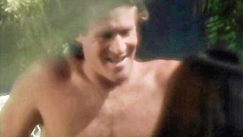 Shirtless curly haired retro hunk smiling while nuzzling brunette woman's neck poolside