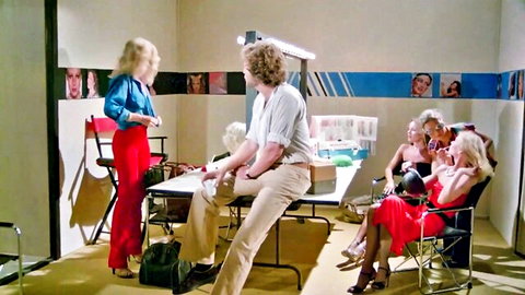 Vintage retro porn casting scene with standing blonde babe in red pants talking to frizzy-haired man at table with seated blonde in red dress