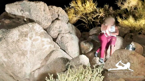 Housewife sits on desert rocks at night with pink leggings down holding pot between spread legs