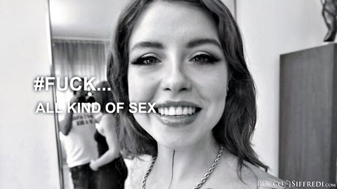 Smiling young brunette with long hair poses topless in mirror as man stands behind in Rocco Siffredi scene