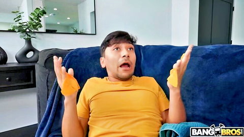 Excited Latino guy in yellow shirt holds orange gloves wide-eyed on blue couch Bang Bros