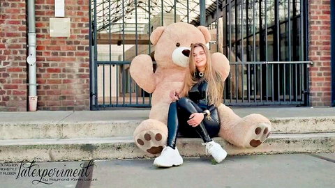 Blonde woman in tight latex outfit hugging giant teddy bear outdoors