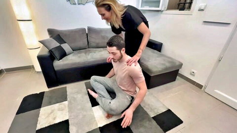 Blonde MILF massages man's shoulders from behind in yoga pose on living room rug