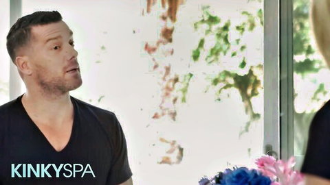 Handsome bearded stud in KinkySpa shirt talks to sexy blonde near window with flowers