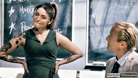 Tattooed brunette teacher in glasses and green dress smirks at suited man in taboo classroom confrontation