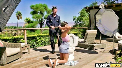 Emily Willis kneeling restrained in short skirt, outdoor BTS for Monsters of Cock