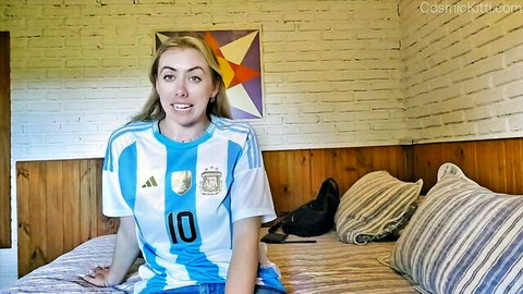 Blonde in Argentina soccer jersey smiling on bed at Uruguay nude retreat