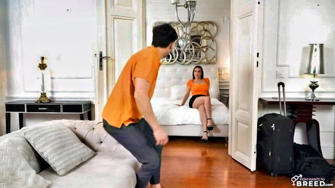 Stepmom in orange top and skirt on bed, man approaches for creampie temptation