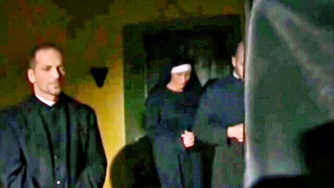 Two priests in black cassocks surround nun in habit in dimly lit retro room