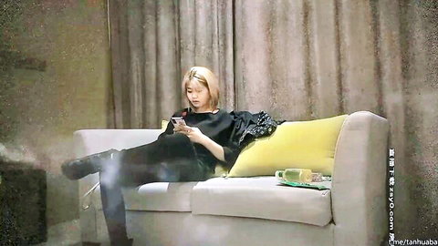 Blonde short-haired Asian woman in black outfit lounging on couch with phone