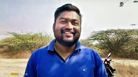 Handsome bearded Indian man smiling outdoors next to motorcycle in dry field