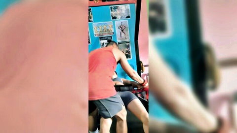 Two muscular men in gym, one in red tank top and shorts bending over suggestively