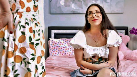 Busty brunette MILF in glasses and cleavage-baring white top sits on pink bed with woman in orange dress