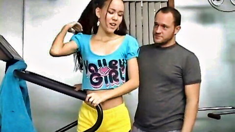 Petite East Asian teen with small tits flexes bicep in tight blue varsity top and yellow shorts at gym with older man