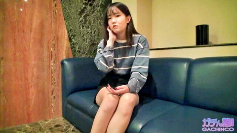 Cute Asian amateur girl in striped sweater and short skirt sitting on blue sofa touching cheek