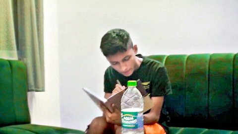 Young Desi boy writing in notebook on couch with water bottle