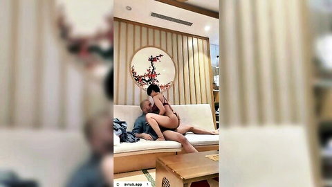 Asian woman in pink top rides man's lap intimately on couch